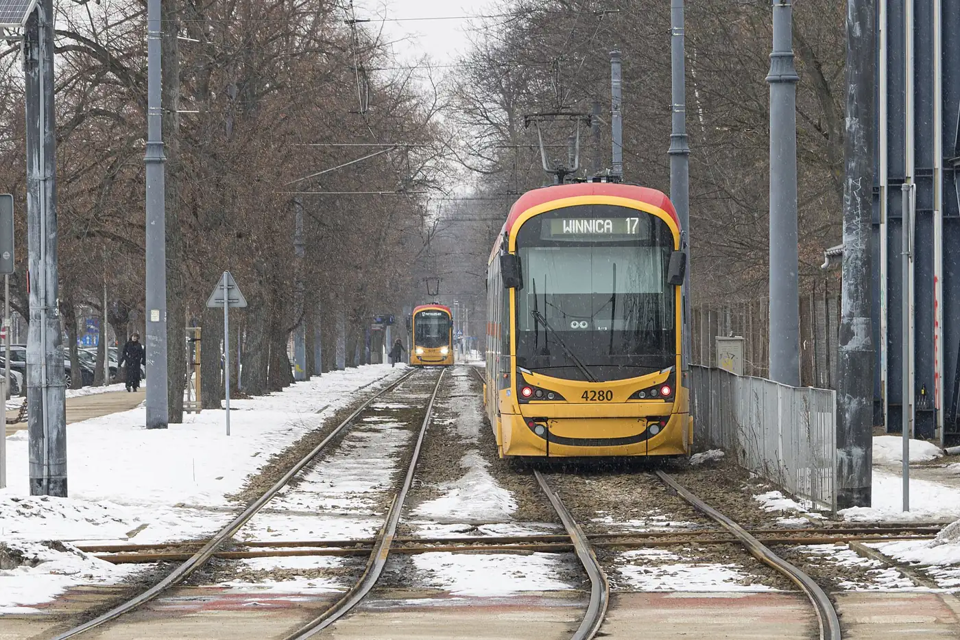 A yellow and red Warsaw tram approaching along a tree-lined track on a snowy winter day.
