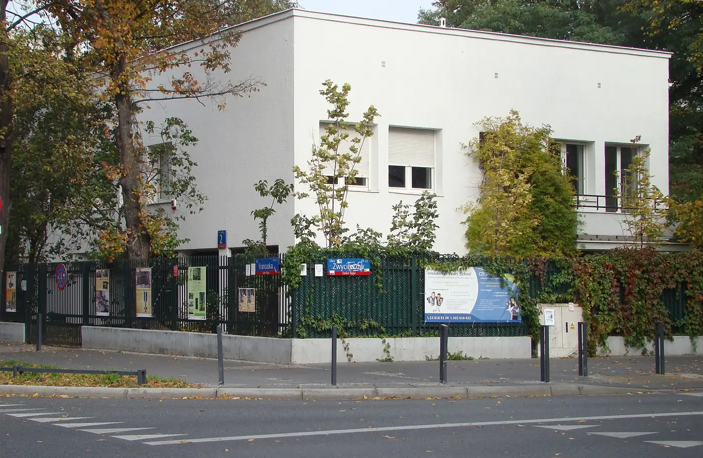 A white 1920s modernist villa behind a fence on a quiet tree-lined street in Saska Kępa.